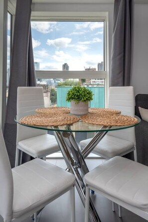Een moderne eetruimte met een glazen tafel, witte stoelen en stadszicht door grote ramen.