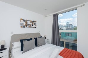 A modern bedroom with a city view, a large bed with multiple pillows, a nightstand, and a unique wall art piece.