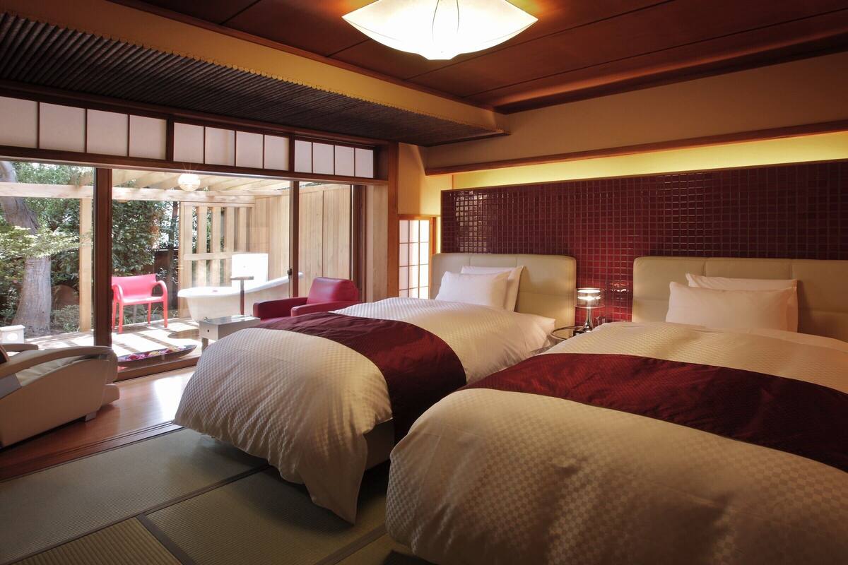 A hotel room with two beds, a red chair, a small table, and a view of the outdoors.