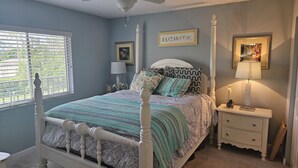 Ein Schlafzimmer mit einem weißen Himmelbett, Deckenventilator, Nachttisch mit Lampe und Fenster mit Jalousien.