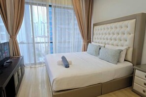 Une chambre d’hôtel avec un grand lit, un téléviseur posé sur un bureau et une vue sur les immeubles à travers des rideaux transparents.