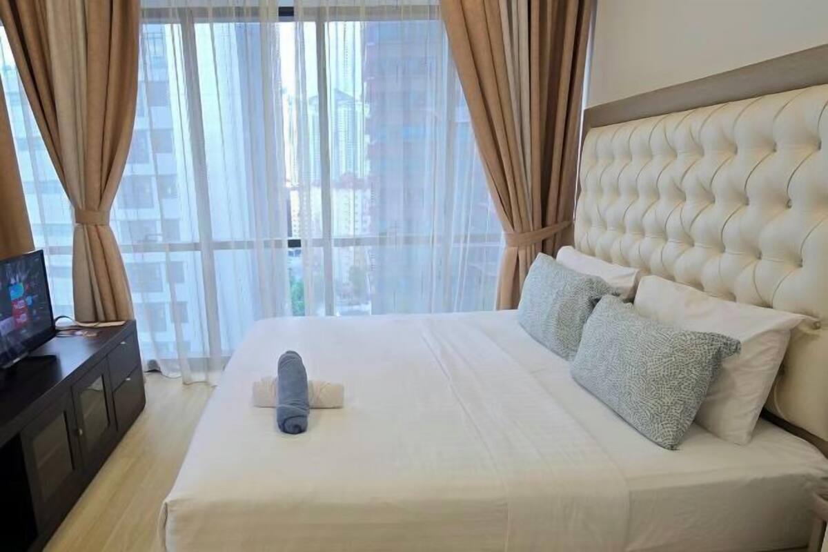 A hotel room with a large bed, a television on a dark wooden side table, and sheer curtains.