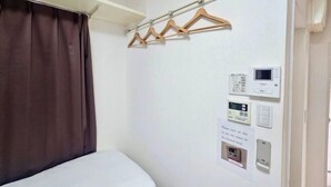 A hotel room with a bed, a curtain, a coat rack with hangers, and a wall-mounted control panel with buttons and a display.