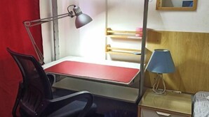 Habitación pequeña con escritorio, silla y cama. Incluye una lámpara de escritorio, una lámpara de noche y un cesto de basura pequeño.