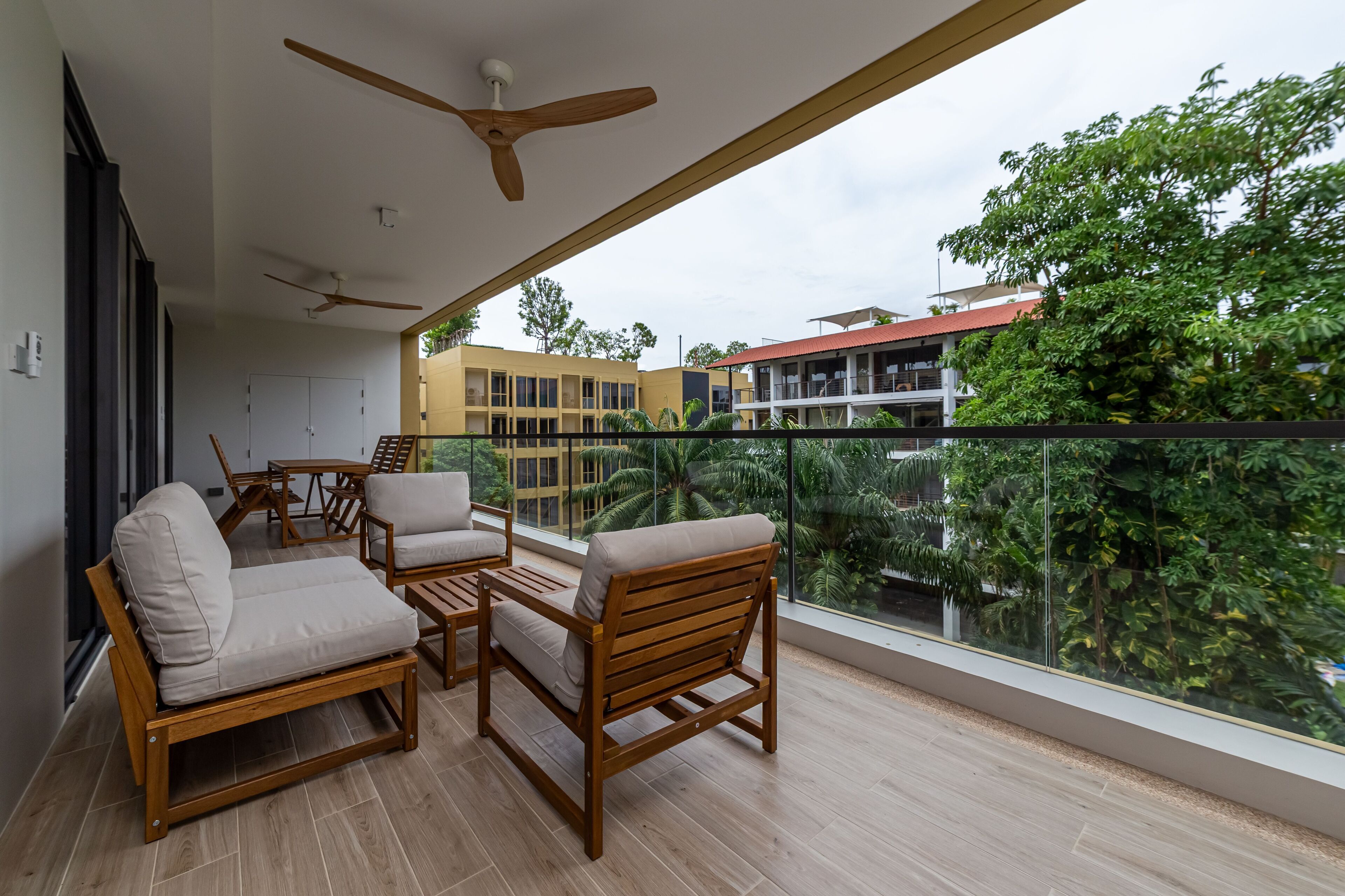 A balcony with wooden furniture and ceiling fans, overlooking a building and lush greenery.