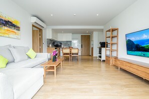 Een moderne woonkamer met een bank, salontafel en een televisie op een houten plank. Achtergrond is een keuken zichtbaar met kasten en een eethoek.