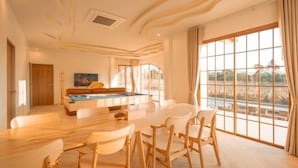 Una habitación amplia con mesa de billar, zona de comedor con sillas y ventanas grandes que ofrecen vistas al exterior.