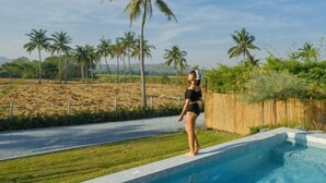 Una persona de pie junto a una piscina, con vistas a palmeras y una valla al fondo.