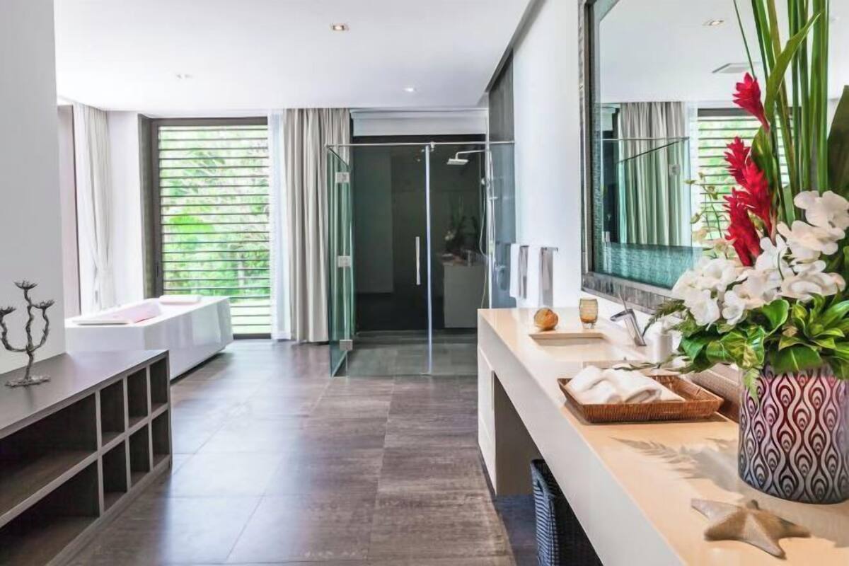 A modern hotel room with a large window, a glass shower, a white sofa, a wooden console table, and a vase of flowers.