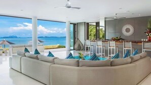 A modern living room with a large sectional sofa, a dining area with white chairs, and a view of the ocean. - Ocean's 11 Villa (Phuket)