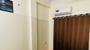 Quarto de hotel com uma cama, um puff marrom, ar-condicionado fixado na parede, câmera de segurança instalada na parede, quadro pendurado na parede e um abajur.
