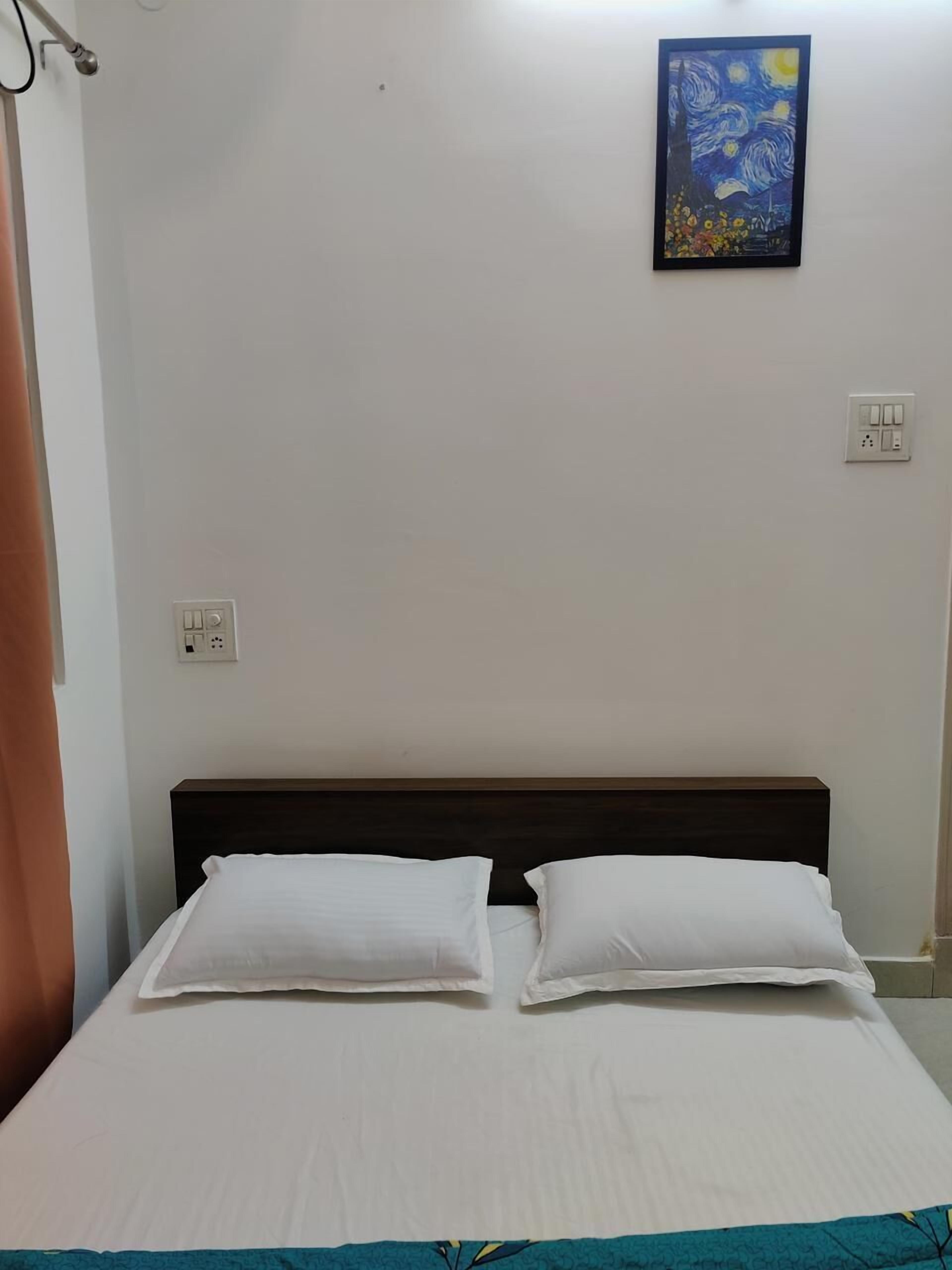 A bedroom with a bed, white pillows, a dark headboard, a painting of a night sky, and a power outlet on the wall.