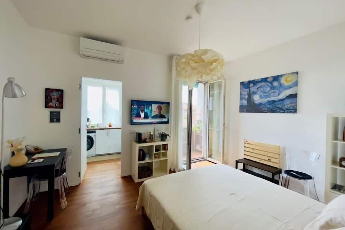 A hotel room with a bed, a desk, a chair, a TV, a painting, and a bench.