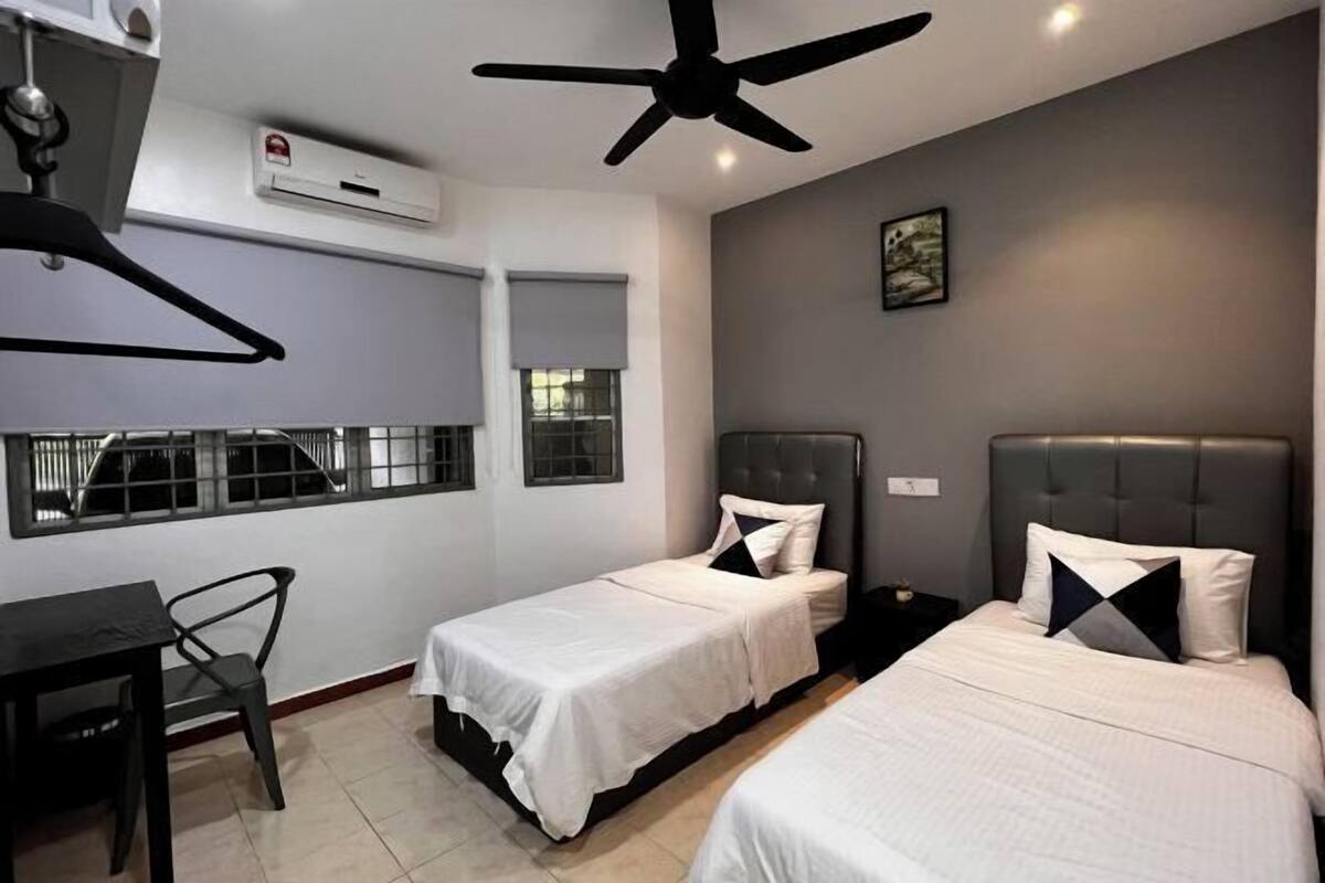 A hotel room with two beds, a desk, and a chair. There is a ceiling fan, an air conditioning unit, and a window with blinds.
