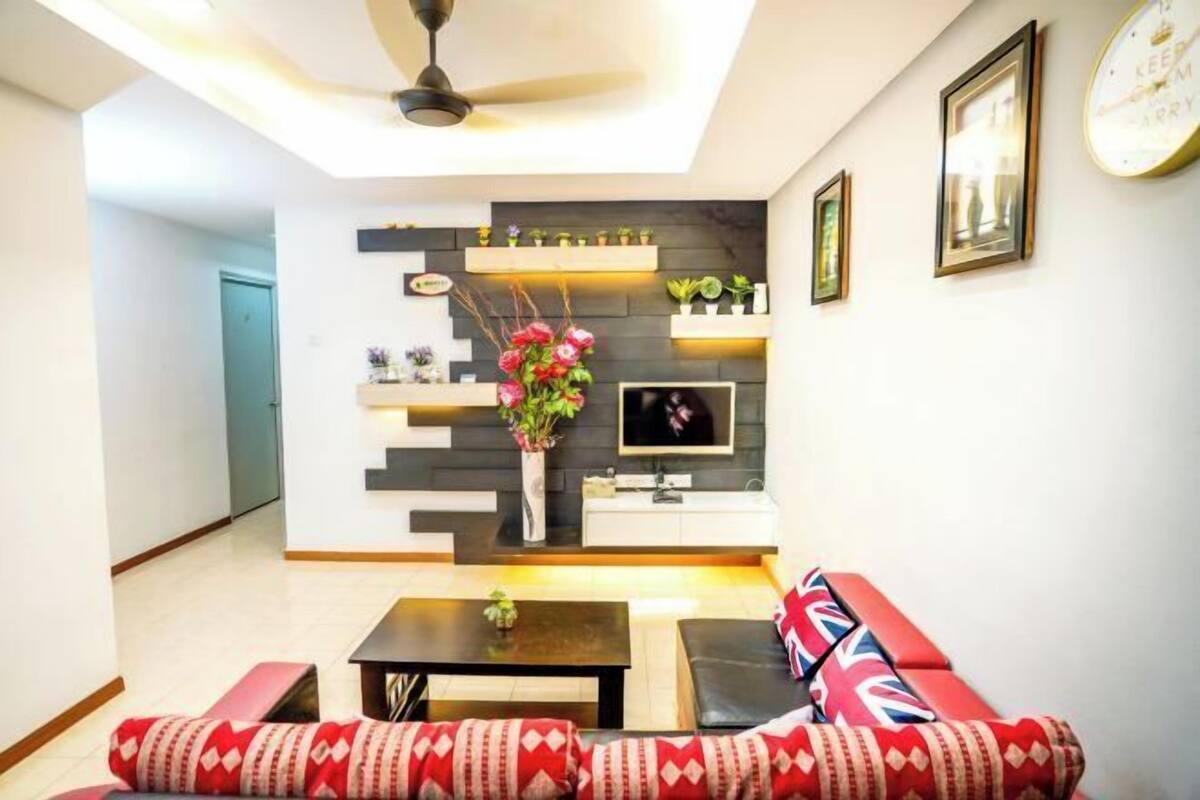 Una sala de estar moderna con un sofá, una mesa de centro y un televisor montado en la pared. Se observan estanterías con objetos decorativos y un ventilador de techo.
