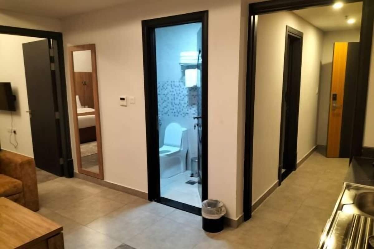 Ein Flur, der zu einem Badezimmer mit Toilette und Waschbecken sowie einem Mülleimer und einer Holztür mit Spiegel führt.