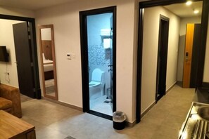 Pasillo que conduce a un baño con inodoro y lavamanos, un cubo de basura y una puerta de madera con espejo.