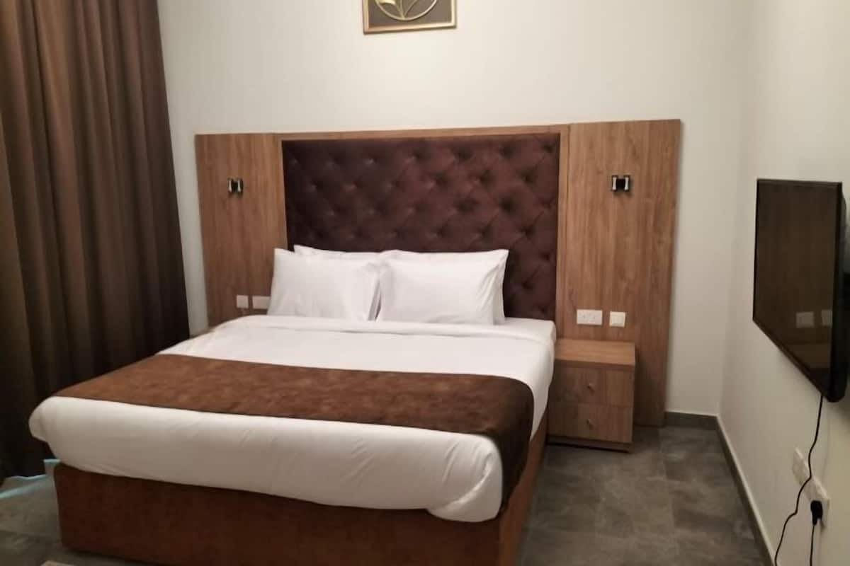 Ein ordentlich gemachtes Bett mit braunem Kopfteil und weißer Bettwäsche, ein kleiner Nachttisch und ein an der Wand montierter Fernseher.