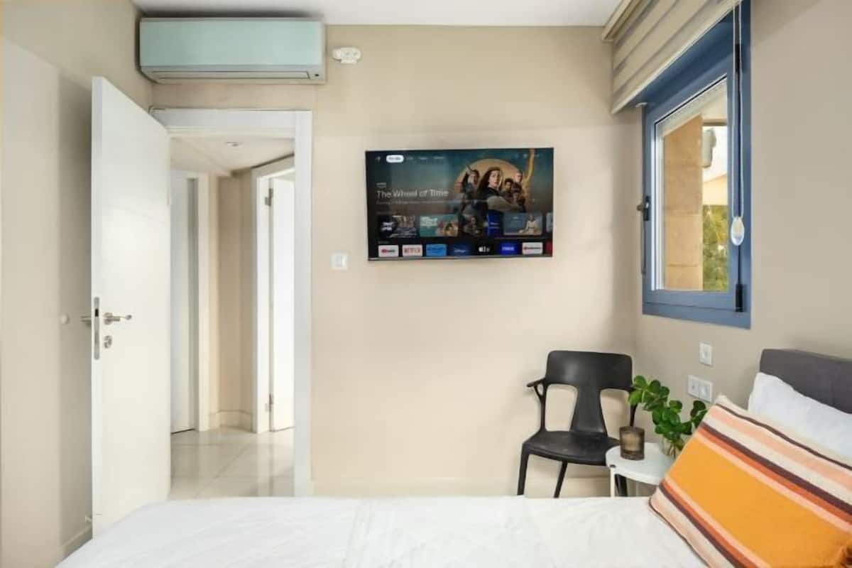 Habitación de hotel con cama, televisión en la pared mostrando un servicio de streaming, una silla, una mesita con una planta y ventana con vistas.