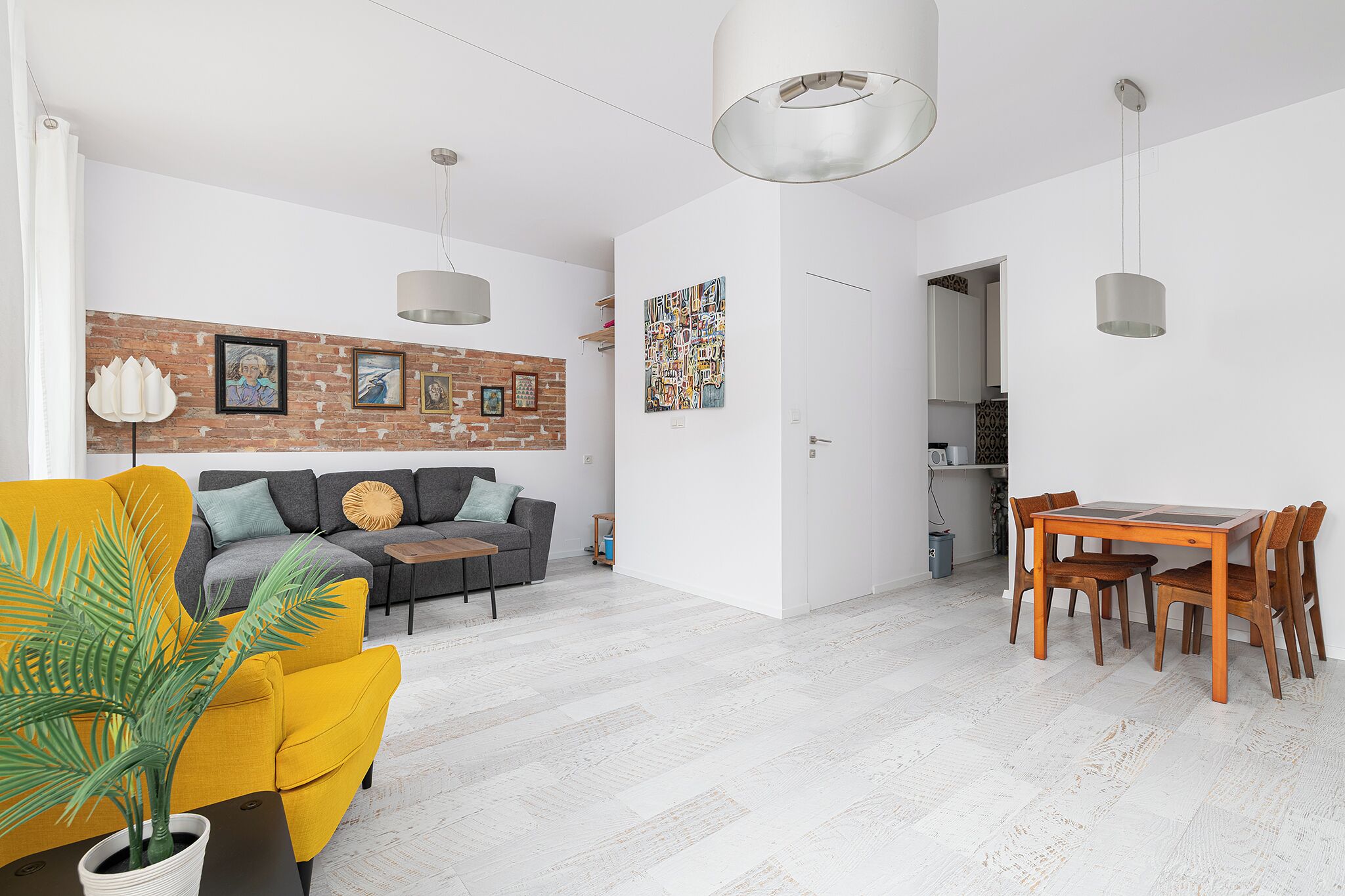 Un salon moderne avec un canapé, une table à manger et des chaises. On y trouve un mur en briques orné de tableaux encadrés et une porte blanche.