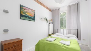A bedroom with a bed, a wooden dresser, a painting, and a window with curtains. - RentPlanet - Apartament Mirów II (Warsaw)