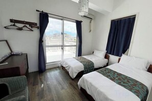 A hotel room with two beds, a desk, a chair, and a view of the city through a large window. - Yonna Yonna よん  なよんな (Miyakojima)