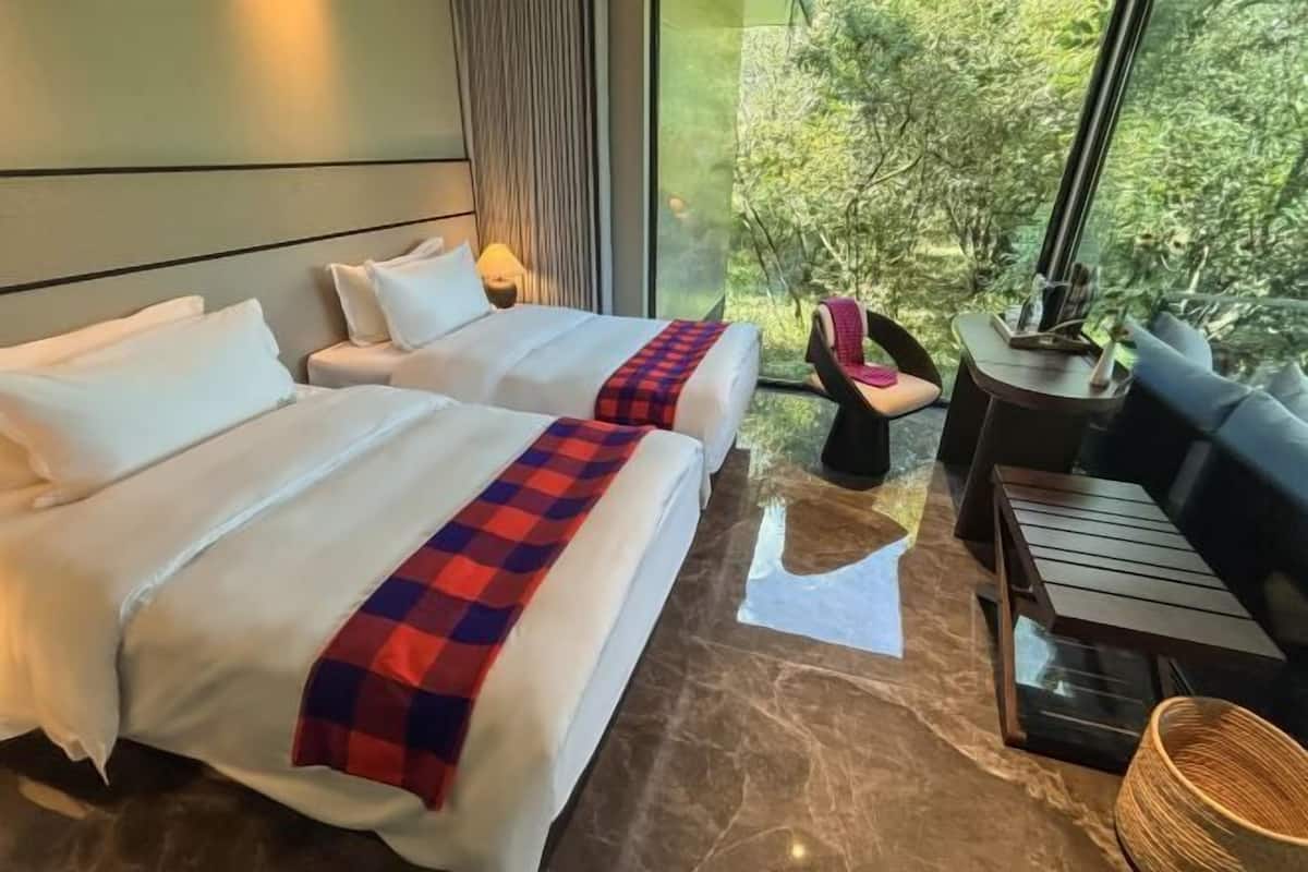 A hotel room with two beds, a sofa, a small table, and a view of greenery outside.