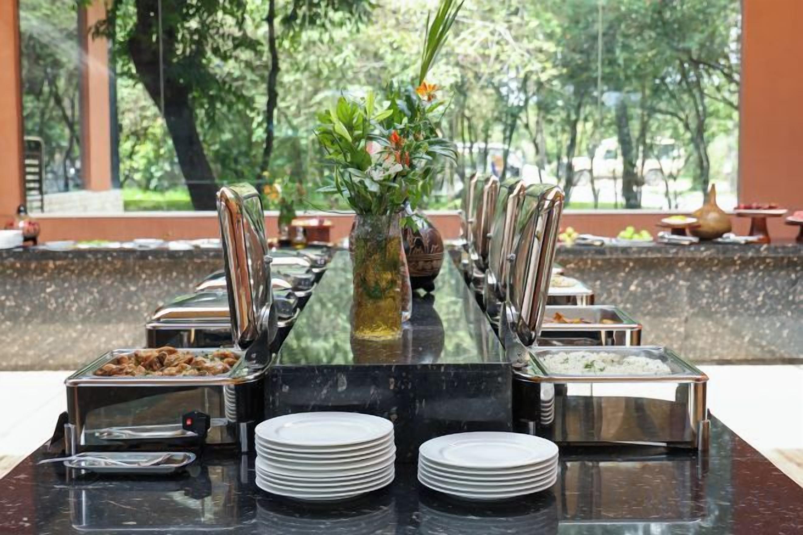 A buffet setup with multiple serving stations, a central vase with flowers, and a view of a green outdoor area.