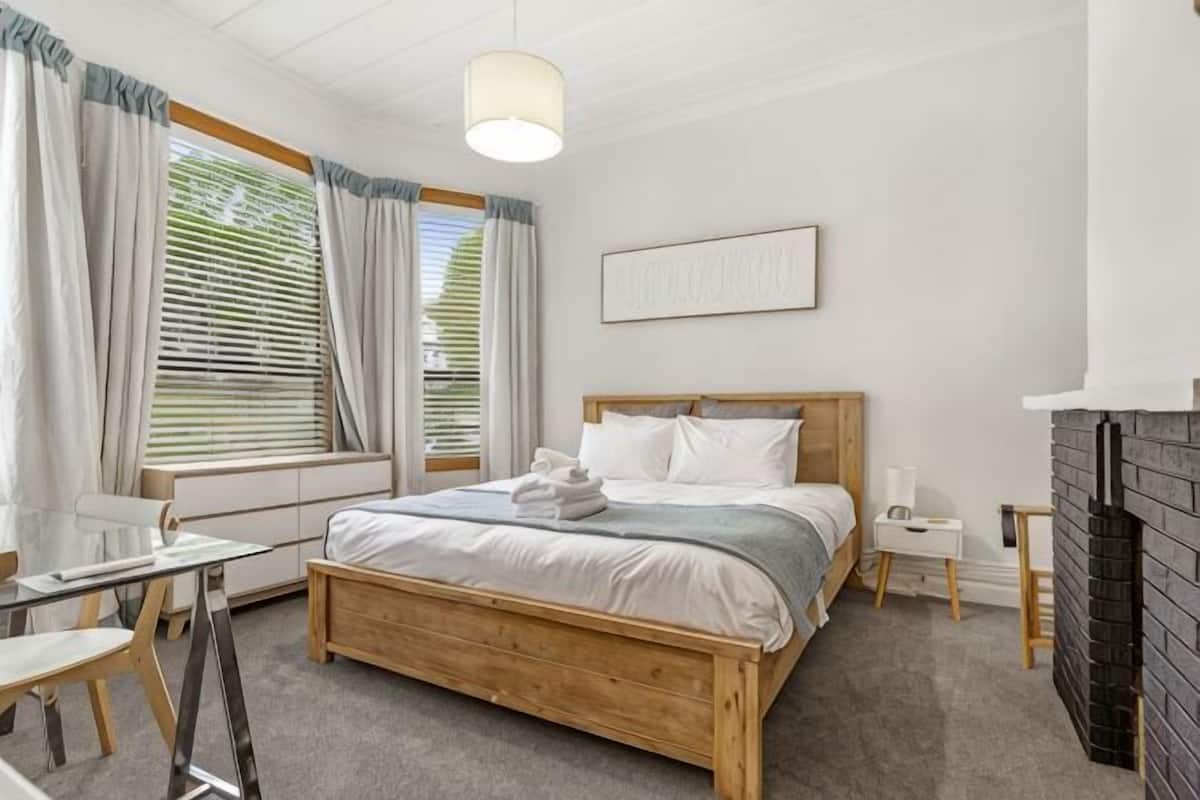 Ein Schlafzimmer mit einem hölzernen Bett, einem Nachttisch, einem Sessel, einem Schreibtisch und einem Kamin.