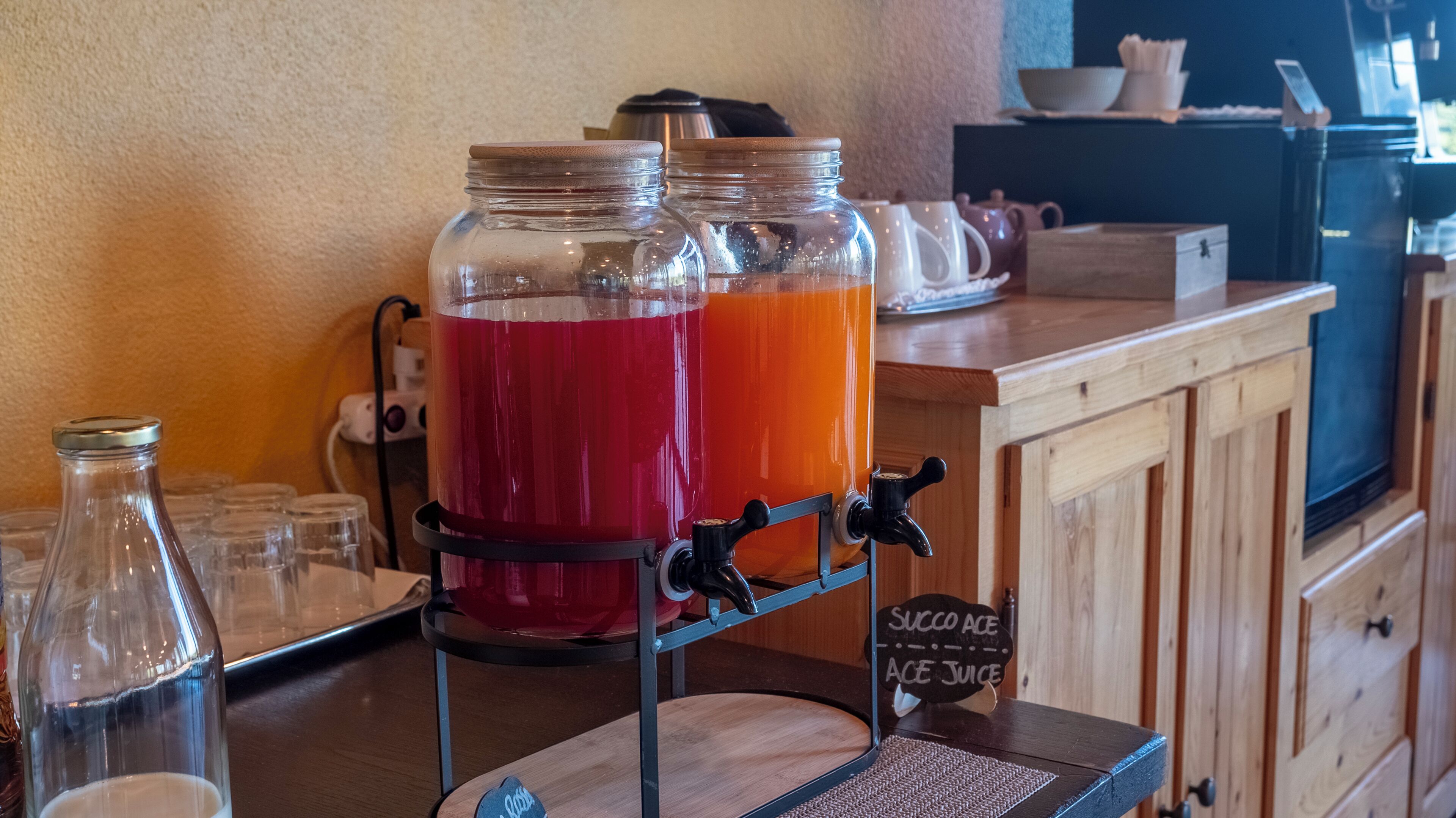 Una estación de bebidas con dos jarras grandes de jugo, un letrero que dice 'SUCCO ACE JUICE' y un mueble de madera al fondo.