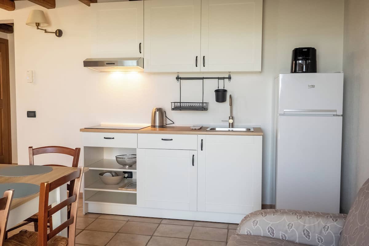 A compact kitchen with white cabinets, a stainless steel kettle, a coffee maker, and a refrigerator.