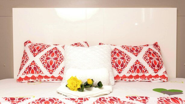 A bed with a white pillow and two red and white patterned pillows, a yellow rose, and a red and white patterned bedspread. - AM Appart's Suite Jacuzzi de luxe in Luanda (Kilamba)