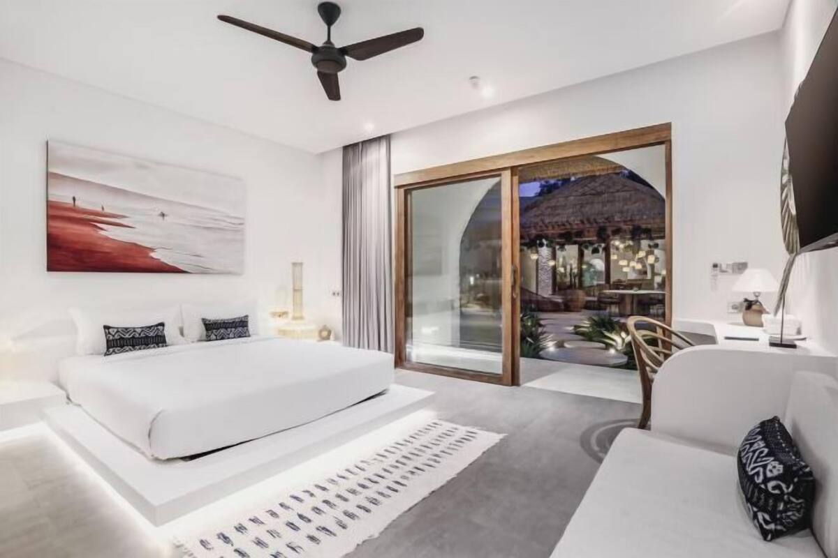 Une chambre moderne avec un grand lit, un ventilateur de plafond, une vue sur la plage par une porte-fenêtre coulissante et une bibliothèque.