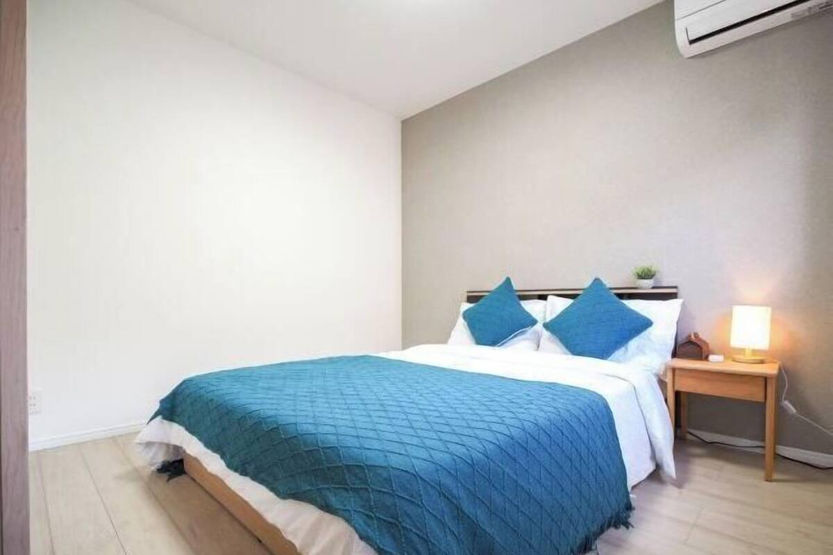 A neatly made bed with a blue bedspread and pillows, a bedside table with a lamp, and an air conditioning unit on the wall.