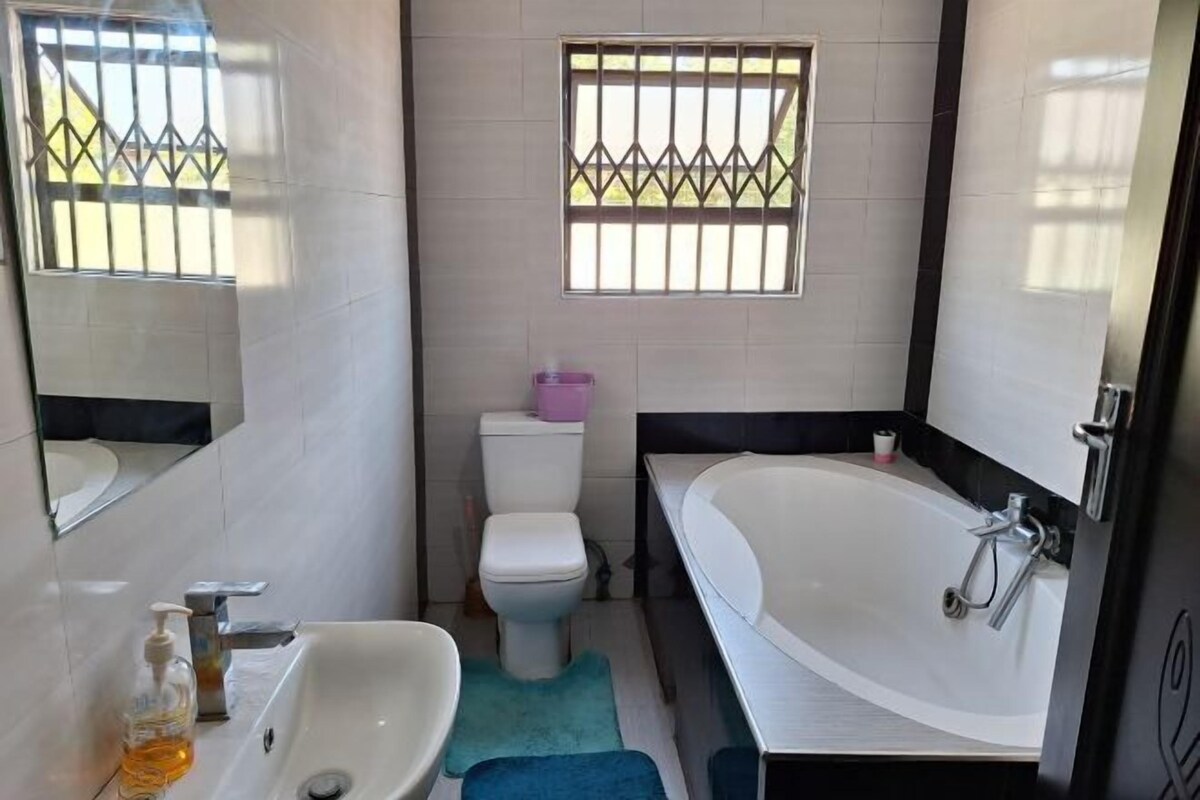 Une salle de bain avec un lavabo, des toilettes et une baignoire, tous de couleur blanche. On y trouve une fenêtre avec des barreaux, un miroir et une poubelle rose.