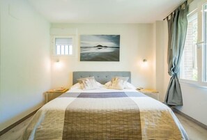 Un dormitorio con cama, un cuadro, una ventana con cortinas y una mesita de noche.