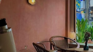 A cozy indoor seating area with wicker chairs and a round table, a decorative wall light, and a view of a street with greenery outside. - DEJA HOME (Chiang Mai)