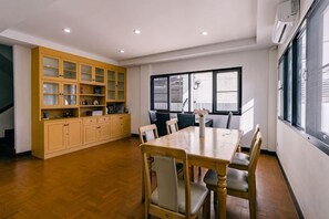 A dining area with a wooden table and chairs, a kitchen with open shelving, and a large window providing natural light. - DEJA HOME (Chiang Mai)