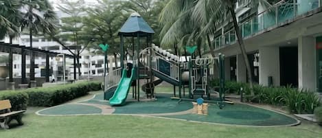 A playground with a slide and climbing structures, surrounded by a green lawn and palm trees.
