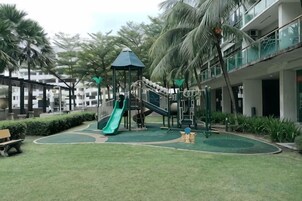 A playground with a slide and climbing structures, surrounded by a green lawn and palm trees.
