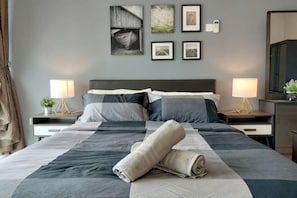 Ein ordentlich bezogenes Bett mit einem zusammengerollten Handtuch, zwei Nachttischlampen und gerahmten Bildern an der Wand.
