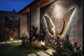 A modern building exterior with a concrete wall, a wooden door, and a large illuminated wing-like structure. - Villa Palm Angels by BaliSuperHost (Bali)