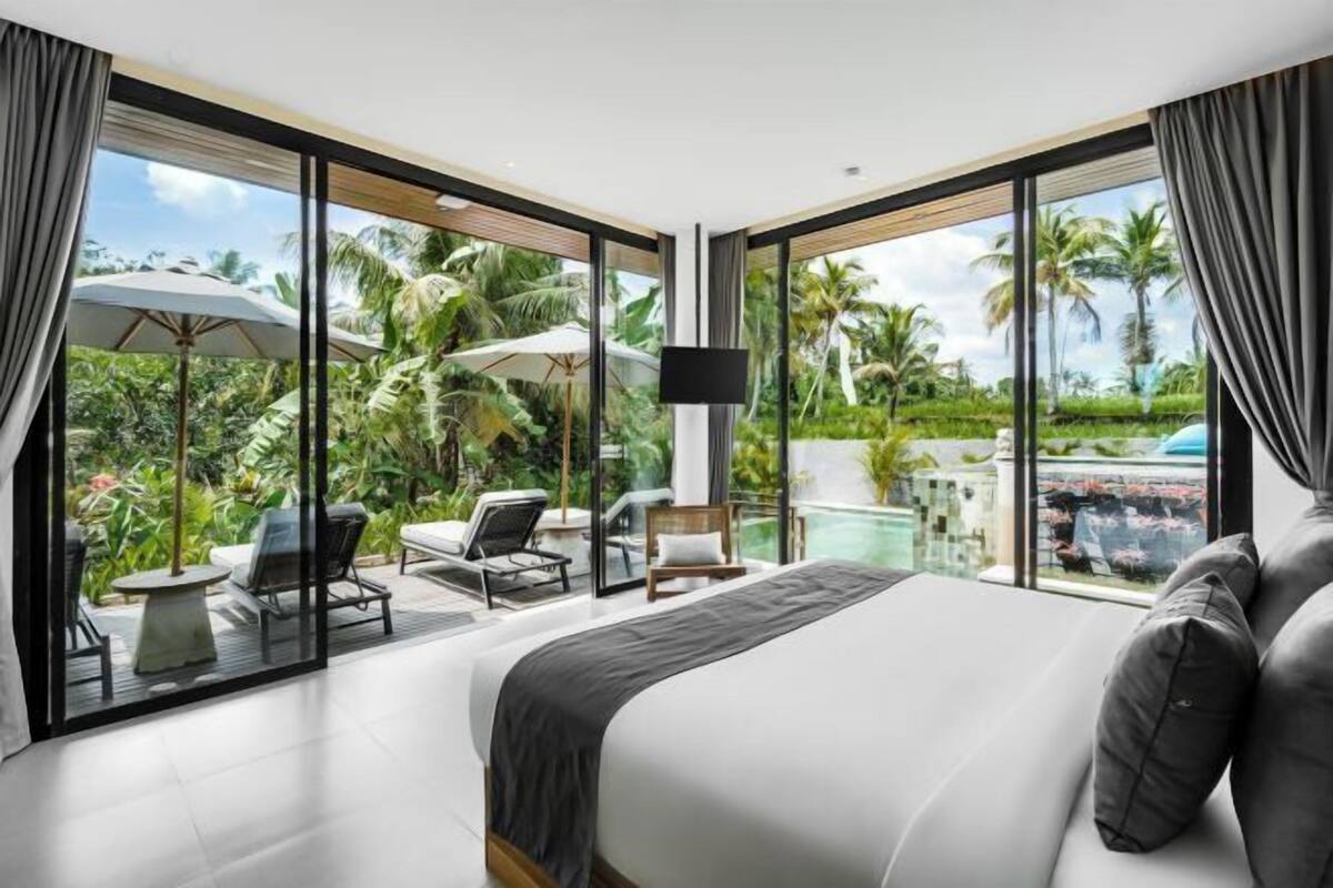 A modern hotel room with a large bed, a TV, and a view of a tropical garden and pool.