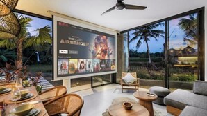 A modern interior with a large screen displaying a Netflix movie, a dining area with a table set for two, and a seating area with a sofa and a small round table. - Villa Palm Angels by BaliSuperHost (Bali)