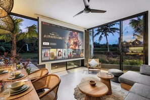 A modern living room with a large flat-screen TV displaying a Netflix show, comfortable seating, and a view of the outdoors. - Villa Palm Angels by BaliSuperHost (Bali)