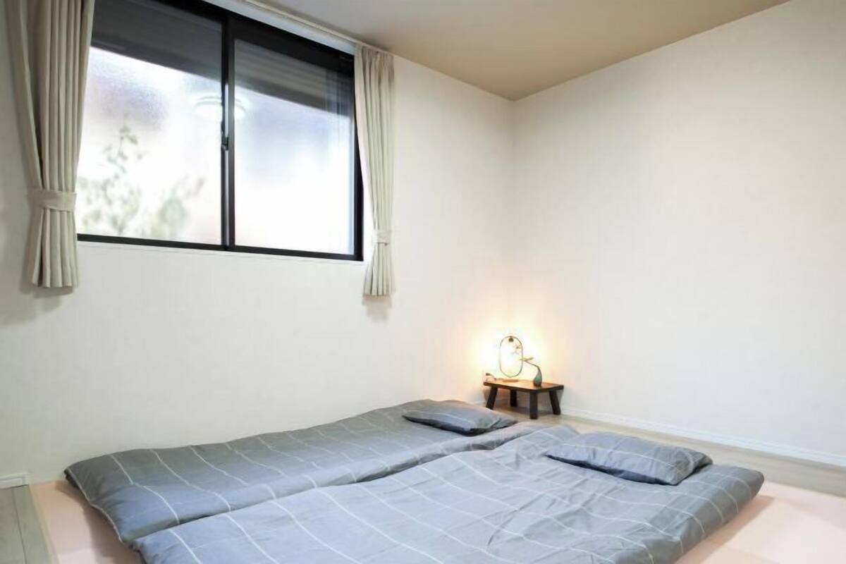 A single bed with a grey bedspread and pillows, a bedside table with a lamp, and a window with curtains.
