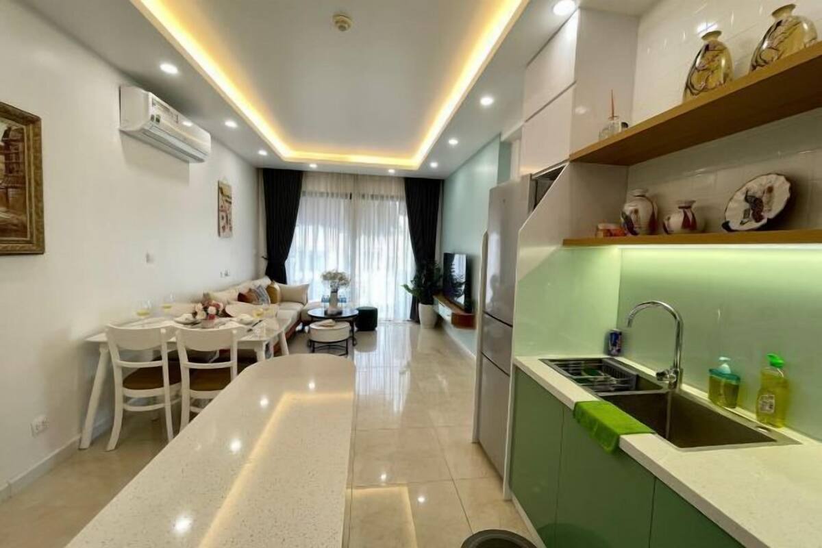 Eine moderne Küche mit einer zentralen Kochinsel, ein Essbereich mit einem weißen Tisch und Stühlen sowie ein Wohnzimmer mit Sofa und Couchtisch.