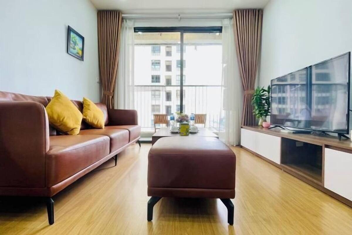 Ein modernes Wohnzimmer mit einem braunen Ledersofa, passendem Hocker, einem Flachbildfernseher auf einem Holzboden und einem großen Fenster mit Vorhängen.