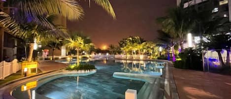 Une piscine éclairée la nuit, entourée de palmiers, avec un bâtiment visible au loin.