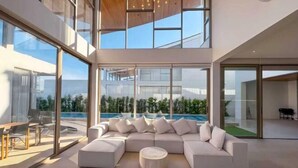 A modern living room with a large sectional sofa, a round coffee table, and a glass wall overlooking a pool. - Trinity Villa 5 Bedroom & 20 Meter Pool Luxury Villa near Bangtao Beach (Phuket)